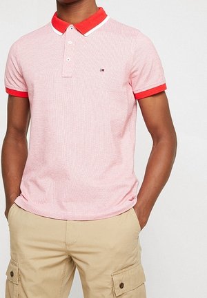 Mann trägt ein kurzärmeliges pinkes Poloshirt mit rotem Kragen und Ärmelabschlüssen, kombiniert mit beigen Cargoshorts, Hände in den Taschen.