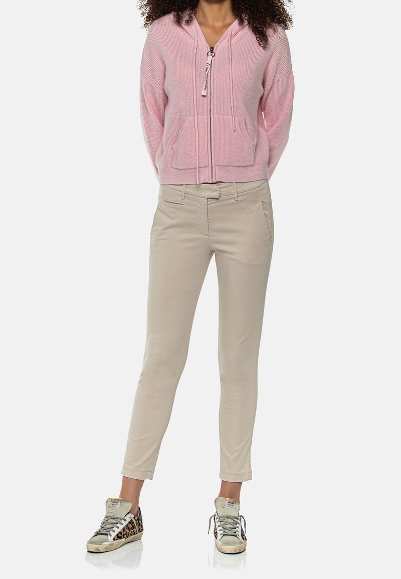 Femme portant un sweat à capuche rose avec fermeture éclair, pantalon beige ajusté et baskets à imprimé léopard, debout les mains derrière le dos.