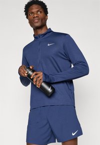 Ναυτικό μπλουζάκι αθλήματος με μακριά μανίκια, μισό φερμουάρ και ανακλαστικό λογότυπο, συνδυασμένο με ναυτικές σορτς. Το μοντέλο κρατάει μια μαύρη αθλητική φιάλη νερού.