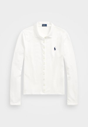 Valge pikakäistega polo särk, milles on nupplist ja pehme tekstuur, ning väike tumesinine logo vasakul rinnal.