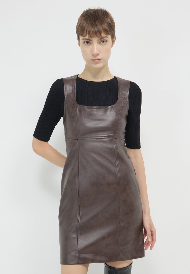 Junge Frau, die ein ärmelloses Kleid aus dunkelbraunem Leder über einem schwarzen, gerippten T-Shirt mit kurzen Ärmeln trägt, steht vor einem schlichten weißen Hintergrund.