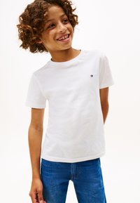 Lächendes Kind mit lockigem Haar, das ein weißes Kurzarmhemd und blaue Jeans trägt und vor einem weißen Hintergrund steht.