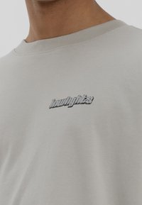 Camiseta de algodón gris claro con un logo brillante y en relieve en negro en el área del pecho izquierdo. Cuello redondo con detalles en canalé.