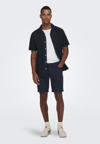 Marineblå kortermet skjorte med knapper over en hvit T-skjorte, kombinert med marineblå shorts. Modellen har på seg kremfargede sneakers og lyse sokker. Enkelt og rent design.
