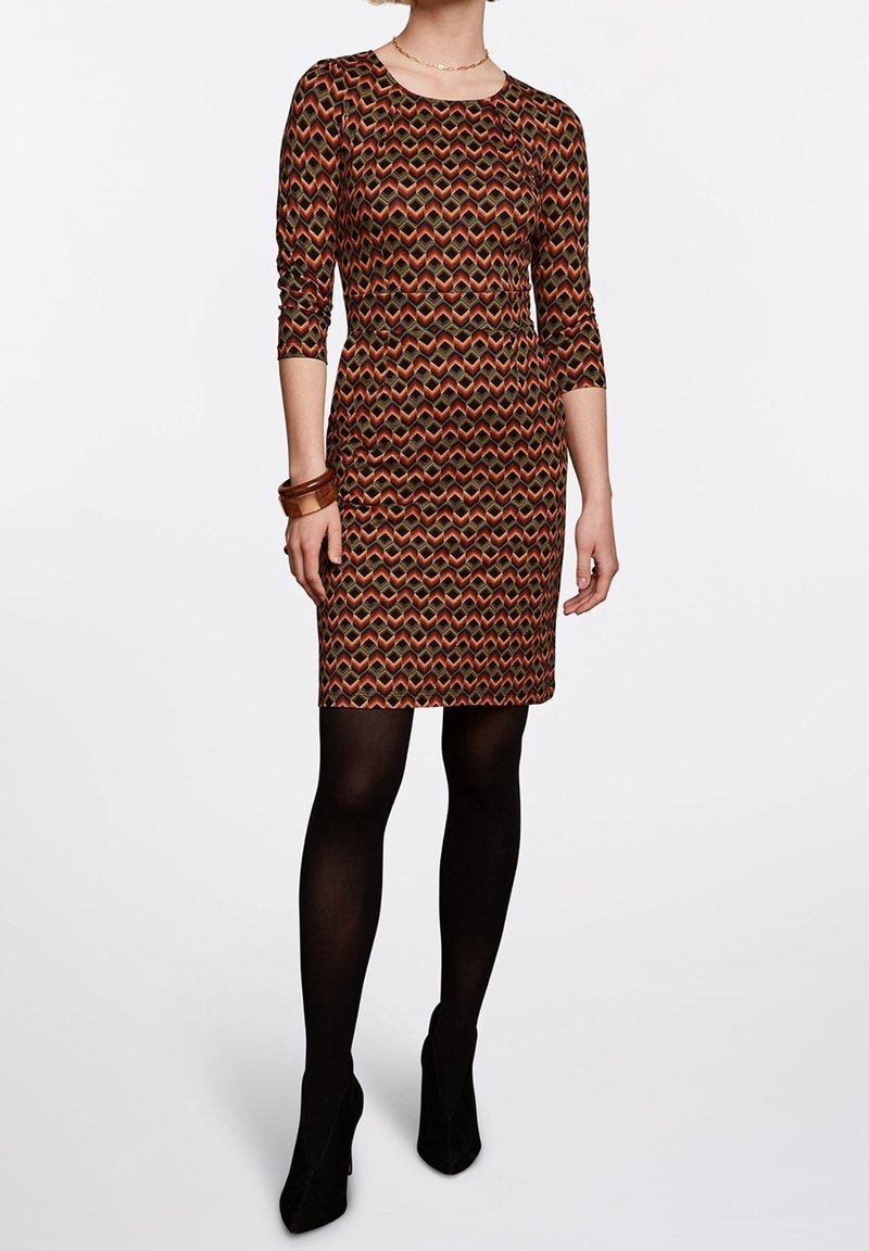 Femme portant une robe ajustée mi-longue aux genoux avec un motif géométrique orange, marron et noir, des collants noirs, des talons noirs et des bracelets en bois.