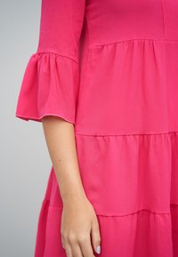 Fuksianvärinen mekko, jossa on levenevät hihat, kerroksittainen malli, teksturoitu kangas ja sauma, joka korostaa vyötärön ja rinnan muotoja.