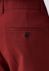 Burgundy színű, testre szabott nadrág, amely karcsú tervezésű, sima tapintású, és hátul húzott zsebe fekete gombbal a biztonságos zárás érdekében.