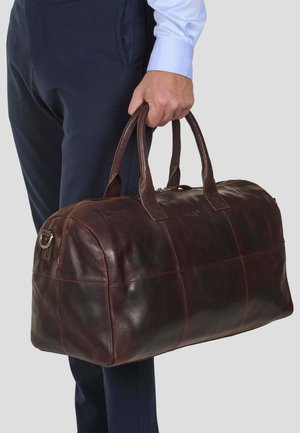 Braune Leder-Duffel-Tasche mit glatter Textur, runder Form, doppelten Griffen und einem Reißverschluss, die von einer Person in blauen Hosen gehalten wird.