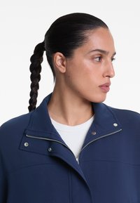 Dunkelblaues Jacket mit hohem Kragen und Reißverschluss, ausgestattet mit metallischen Druckknöpfen und einer glatten, strukturierten Oberfläche. Weißes Hemd darunter sichtbar.