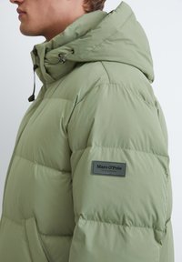 Veste matelassée vert clair, fabriquée à partir d'un tissu lisse et rembourré. Comprend une capuche, une fermeture éclair à l'avant et un logo sur la manche.