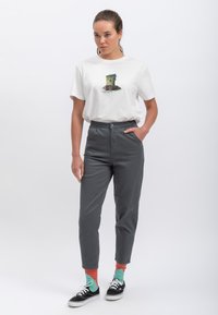 Witte T-shirt met een kleurrijke grafische print, gecombineerd met hoog-waist grijze broek. Model draagt multicolore sokken en zwarte schoenen.