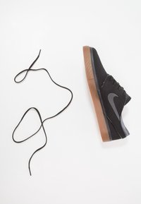 Baskets en toile noire avec semelle marron à côté d'un lacet noir desserré sur fond blanc.