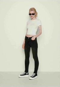 Jauna sieviete, tērpta baltā īsā t-kreklā, melnās pietuvinātās džinsās, melnās augstajās kedās un taisnstūra melnā saulesbrillēs, stāv pret sienu.