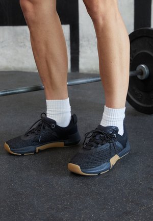 Personne portant des chaussures de sport noires et marron avec des chaussettes blanches debout près d'une barre chargée de poids sur le sol de la salle de sport.