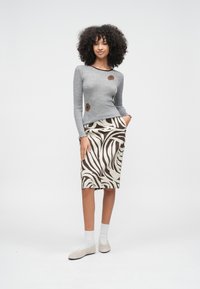 Femeie cu păr creț, purtând un top cu mâneci lungi gri cu pete florale maro și o fustă până la genunchi cu imprimeu zebra, stând cu mâinile în buzunare.