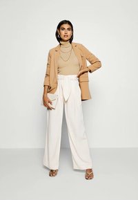 Beige blazer over a fitted beige turtleneck, paired with white wide-leg trousers featuring a tied waist. Accessories include a clutch and strappy sandals.
