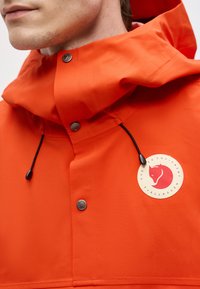 Prim-plan cu o persoană purtând o jachetă cu glugă de culoare portocaliu aprins, cu șnururi negre și logo circular Fjällräven pe piept.