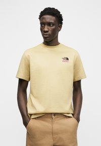 Άνδρας με τα χέρια στις τσέπες, φοράει μπεζ κοντομάνικο πουκάμισο The North Face 1966 και καφέ παντελόνι, στέκεται μπροστά σε λευκό φόντο.