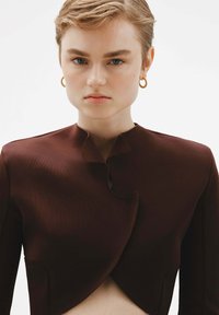 Chaqueta marrón corta con un cuello doblado único, confeccionada en tejido suave. El modelo lleva pendientes de aro dorados, con un fondo neutro.