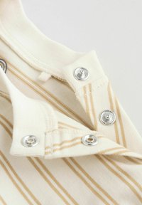 Gros plan sur un tissu à rayures beige avec des boutons-pression métalliques partiellement défaits, montrant les détails de l'encolure et des coutures.