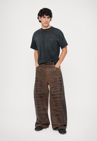 Melns T-krekls ar logotipu, brīvi brūni bikses ar teksturētām akcentiem un plašu piegriezumu, apvienoti ar melnām kurpēm.