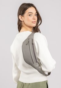 Grijze leren schoudertas met een gebogen vorm, een ritssluitingscompartiment en een verstelbare schouderriem. Gedragen over de schouder, wat goed staat bij een witte trui.