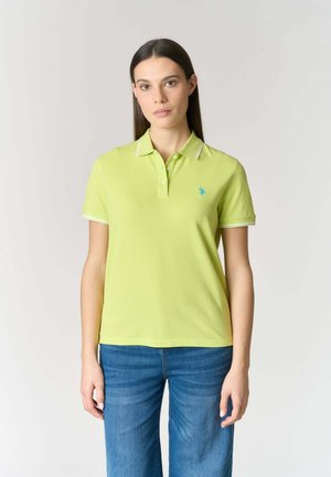 Giovane donna in piedi, rivolta in avanti, che indossa una polo verde chiaro e jeans blu su uno sfondo neutro.