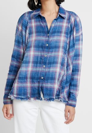 Kvinde iført en blå og lyserød ternet knap-op skjorte med frynser og hvide bukser, stående med afslappede arme ned langs siderne.