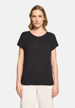 Kvinde med lysbrunt hår iført en enkel sort T-shirt med korte ærmer og off-white bukser foran en ensfarvet lys baggrund.