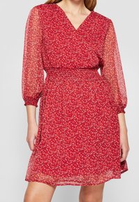 Vestido rojo floral con escote en V, mangas largas transparentes y cintura elástica, llevado por una persona con los brazos relajados.