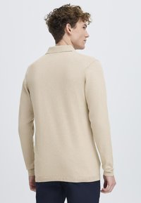 Beige pulóver hosszú ujjakkal, bordázott mandzsettával és gallérral. Kötött textúra, egyenes alsó szegély és diszkrét varrási részletek a vállvarratokon.