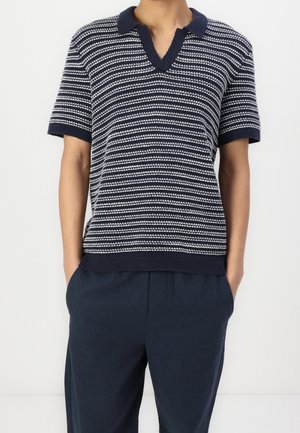 Persoon in een marineblauwe en witte gestreepte korte mouw gebreide polo met donker marineblauwe broek, handen in de zakken, tegen een effen witte achtergrond.