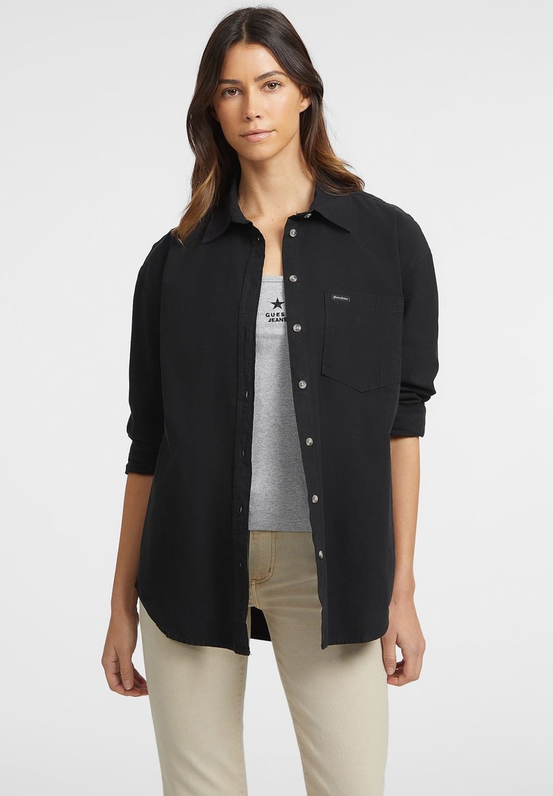 Donna con lunghi capelli scuri che indossa una camicia nera con bottoni sopra un top grigio e pantaloni beige, in piedi contro uno sfondo bianco.