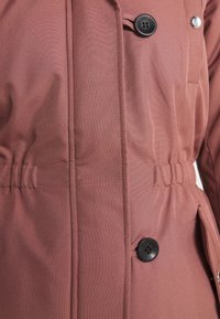 Chaqueta exterior ligera de color marrón rojizo con una superficie texturizada, presenta un cierre de botones, cintura ceñida y bolsillos laterales.