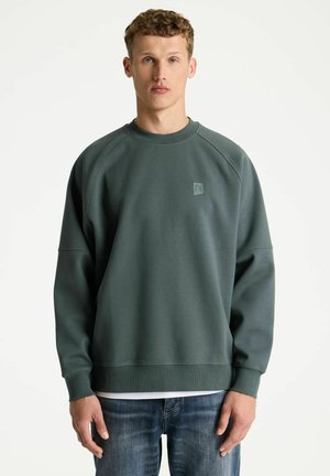 Jonge man met krullend haar die een donkergroene longsleeve trui en blauwe jeans draagt tegen een effen lichte achtergrond.