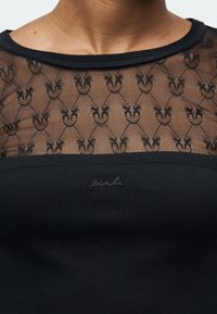 Κοντινό πλάνο ενός ατόμου που φοράει μαύρο τοπ με διάφανο πάνω μέρος από δαντέλα που έχει επαναλαμβανόμενο μοτίβο πεταλούδας και ραβδωτό ύφασμα κάτω από αυτό.