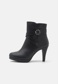 Bota negra de tobillo hecha de cuero sintético con punta afilada, tacón alto y correa decorativa con un detalle metálico circular.