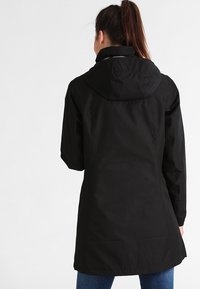 Žena s hnědým culíkem, oblékající dlouhý černý kabát s kapucí a modré džíny, stojící otočená zády před jednobarevným bílým pozadím.