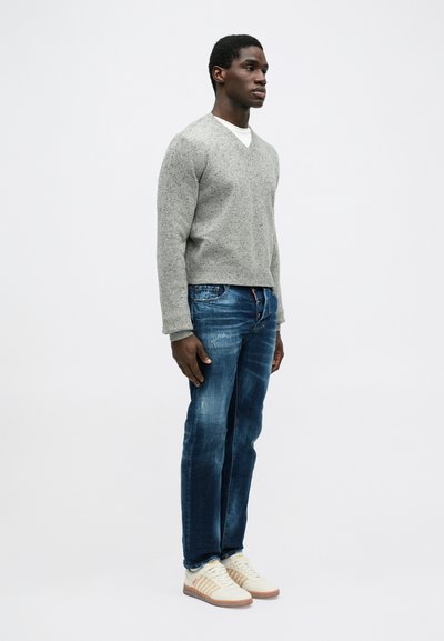 Grijze gebreide V-hals trui, blauwe distressed jeans en witte sneakers met tan accenten. Model staat zijwaarts tegen een neutrale achtergrond.