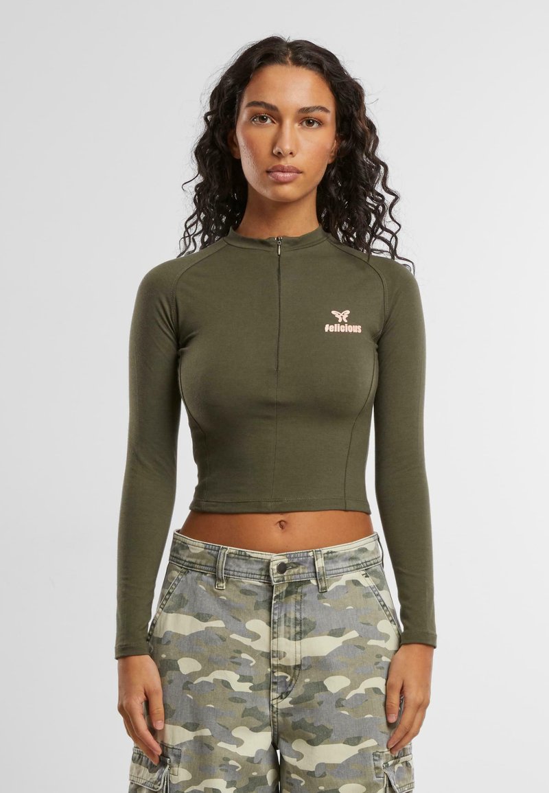 Olivinvihreä pitkähihainen crop-top, jossa on korkea kaulus ja edessä vetoketju. Vasemmassa rinnassa on pieni vaaleanpunainen logo. Sileä pinta.