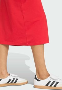 Biele tenisky s čiernymi akcentmi a tromi čiernymi pruhmi, s okrúhlou špičkou a textúrovanou gumovou podrážkou, kombinované s červenou sukňou.