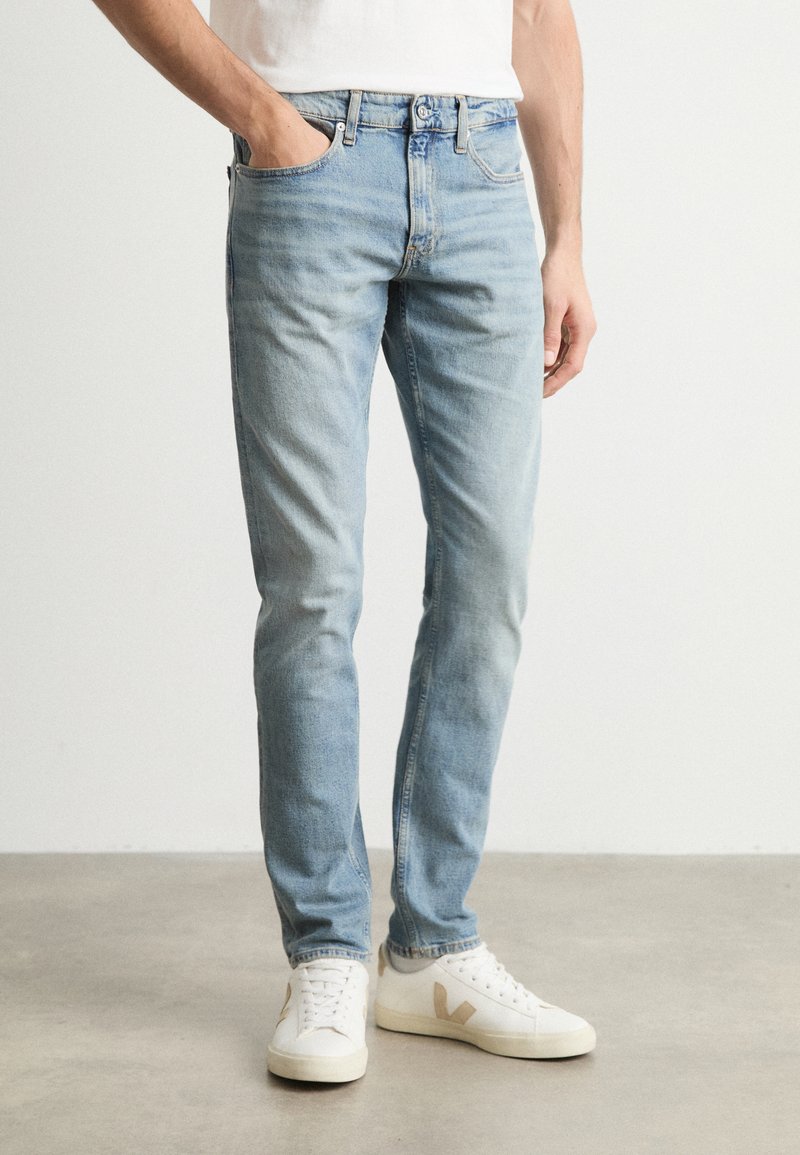 Világoskék, slim fit farmernadrág, öt zsebbel és enyhe kopással. Fehér sportcipőkkel párosítva, bézs kiegészítőkkel.