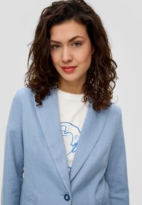 Jasnoniebieska marynarka z klapkami, jednoprzyciskowym zapięciem i teksturowanym materiałem. Noszona na białym t-shircie z niebieskim nadrukiem.