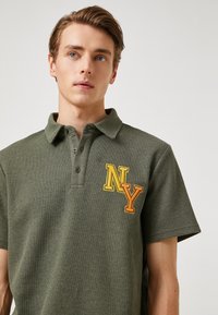 Koszulka polo w oliwkowym kolorze, z teksturowanej tkaniny, z kołnierzykiem, trzema guzikami i haftem "NY" w kolorze pomarańczowym i żółtym po lewej stronie klatki piersiowej.