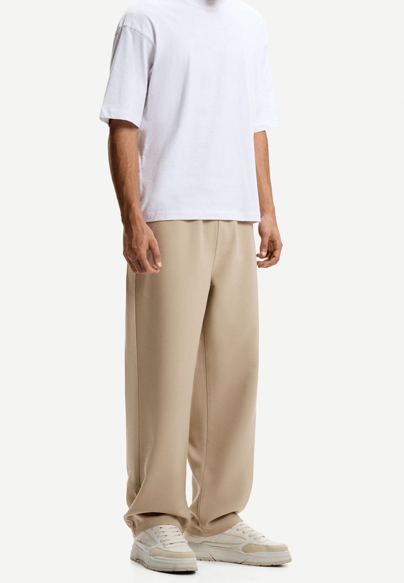 Personne portant une chemise blanche ample à manches courtes, un pantalon beige large et des baskets blanches sur un fond uni.