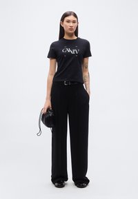 Μαύρο βαμβακερό μπλουζάκι με λευκό λογότυπο, σε συνδυασμό με μαύρο παντελόνι ψηλόμεσο. Αξεσουάρ περιλαμβάνουν μαύρη τσάντα χειρός και γυαλιστερά παπούτσια.