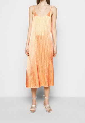 Kobieta ubrana w pomarańczową sukienkę midi na ramiączkach w wzory, z cienkimi ramiączkami, oraz białe sandały na obcasie z paskami.