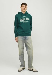 Grøn bomulds-hættetrøje med et hvidt grafisk print, kombineret med lyseblå jeans og beige sneakers med sorte striber. Ensartet baggrund.