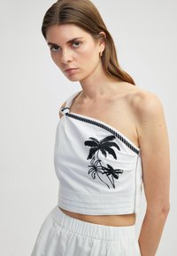 Korte witte top met zwarte palmblad borduursel, een gestructureerde zoom en een zijrits benadrukt door een gedraaide zwarte koordband.