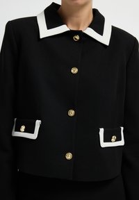 Schwarze, kurz geschnittene Jacke mit einem weißen Kragen, goldfarbenen Knopfverschlüssen und zwei Fronttaschen mit weißen Akzenten. Glatte Stoffoberfläche.